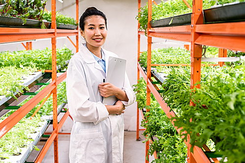 Young successful female researcher or agronomist in whitecoat standing in aisle between shelves with green seedlings