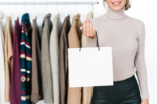 Stylish young woman in light grey sweater and black leather pants holding white paperbag on background of new collection of clothes