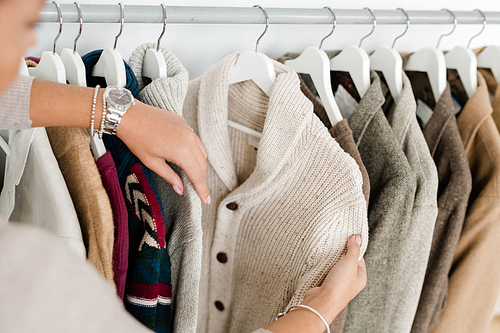 Hands of young woman holding white knitted cardigan while choosing new clothes in modern boutique