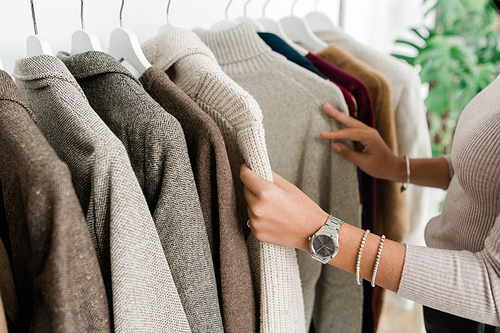 Hands of young female shopper in grey pullover looking through new seasonal collection of casualwear in boutique