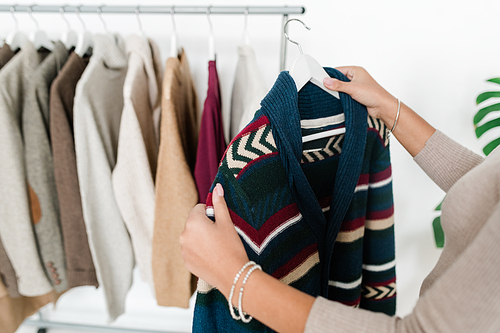 Young woman holding hanger with new warm knitted cardigan while choosing clothes from seasonal collection