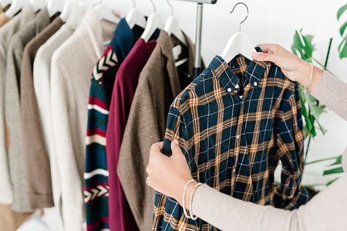 Young female shopper holding hanger with checkered shirt while choosing new casual clothes in boutique