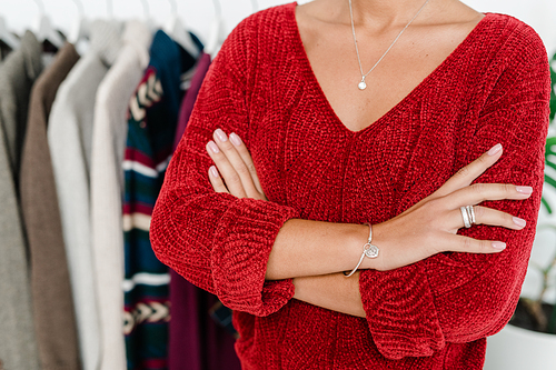 Young elegant woman in red knitted pullover crossing arms by chest on background of rack with new casualwear collection