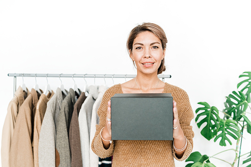 Happy young stylish female in casual pullover holding black box by her chest while standing on background of rack with clothes