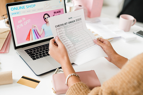 Young female customer holding and reading checklist paper over laptop keypad while sitting by desk