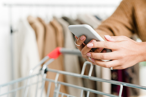 Female shopper hands holding smartphone while scrolling and pushing cart during visit to boutique within seasonal sale period