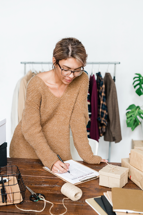 Young casual woman standing by wooden table and checking list of ordered items after receiving parcel from online shop