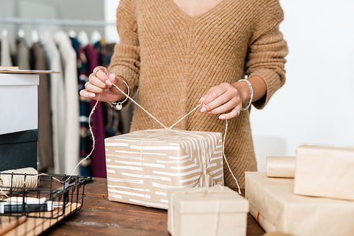 Young casual woman in knitted pullover binding thread on top of wrapped giftbox while packing presents for holiday