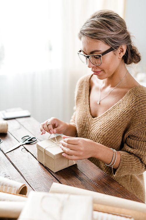 Young female in casualwear and eyeglasses binding wrapped giftbox with threads while sitting by wooden table
