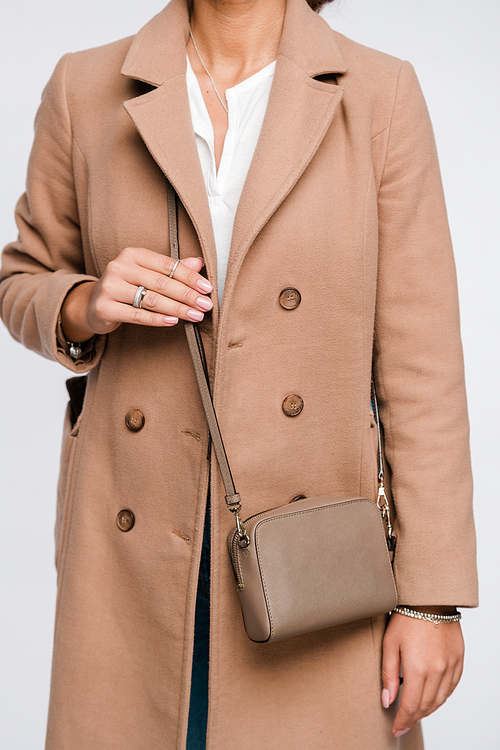 Mid-section of young stylish woman in elegant beige coat with small leather handbag standing in isolation