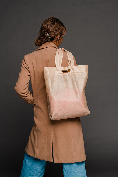 Rear view of young casual woman in elegant coat and blue jeans holding textile tote bag on shoulder