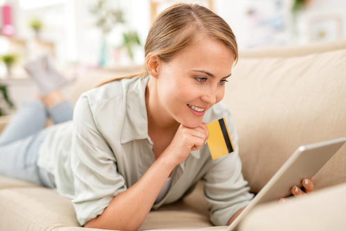 Happy young female with plastic card and touchpad lying on couch and scrolling through online goods while choosing something to buy