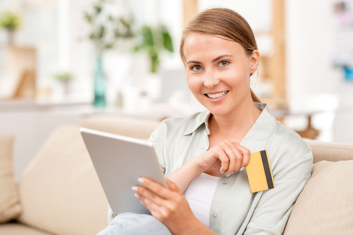 Young female customer using touchpad for searching online goods and plastic card for payment while relaxing on couch
