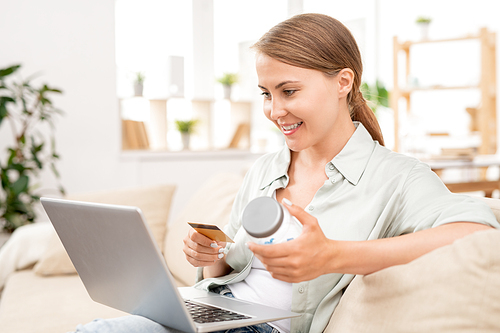 Pretty young woman with plastic card and jar looking at laptop display while going to make order in online shop