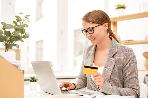 Young successful businesswoman with plastic card looking at laptop display while searching through online goods