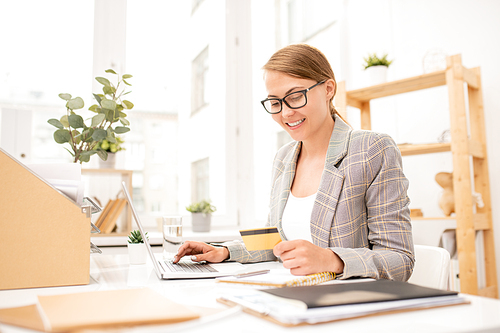 Pretty young office worker looking at personal data on credit card while making online order by workplace