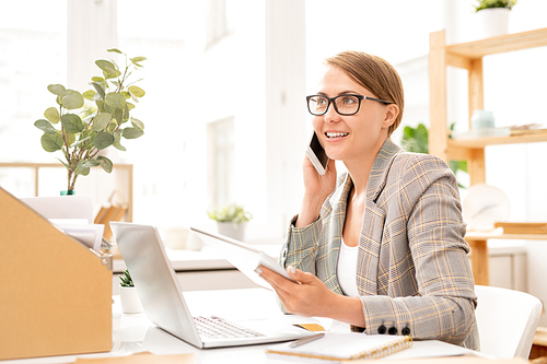 Happy businesswoman talking by smartphone and using tablet in front of laptop while sitting by desk in office