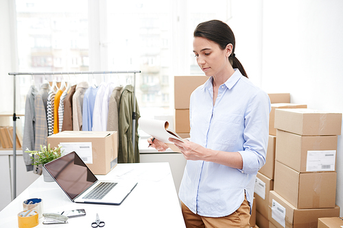 Young female manager reading notes in notepad while preparing goods for packing by workplace