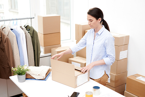 Young office manager in casualwear standing by desk while packing box with online order of client