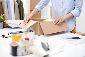 Young manager of online shop office opening carton box by workplace before packing order of client