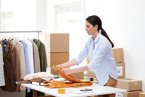 Young casual woman folding yellow velvet pants on desk while preparing order of online shop client