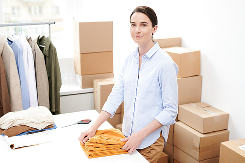 Young brunette female in casualwear looking at you while folding torusers for packing by desk