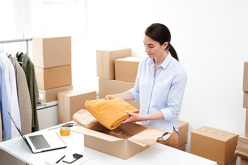 young female online shop manager putting folded velvet yellow pants into box while packing order of client