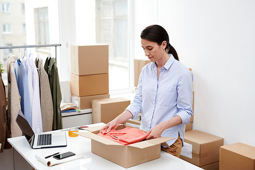 Young attractive woman in casualwear putting folded red pullover into box while packing order of client of online shop