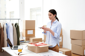 Young female manager with mobile phone photographing packed clothes in open box before sending the order to client