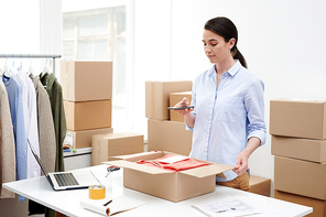 Young female shop-assistant photographing parcel with folded casualwear before packing the box and sending it to client