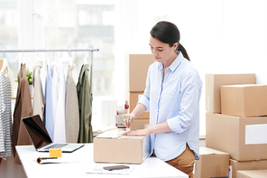 Young online shop manager with adhesive tape dispenser packing carton box with order of client in office