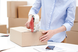 Young female in light blue casual shirt packing box with order of client and sticking it with adhesive tape