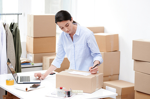 Young female manager of online shop writing down address of client on packed box with order