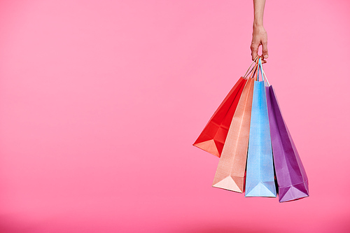 Hand of shopper holding bunch of colorful paperbags or carrying them after shopping on pink background