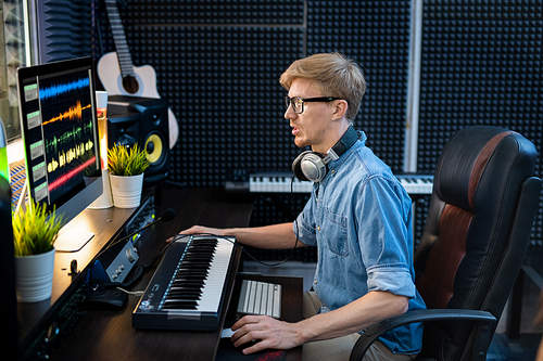 Young male deejay in denim shirt looking at sound mixing tracls on computer screen while sitting by desk in front of monitor
