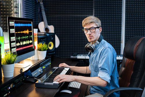 Young casual man with headphones looking at you while sitting by workplace in recording studio in front of computer screen