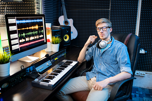 Serious young man in casualwear sitting in armchair by desk with computer monitor and keyboard while mixing sounds in studio