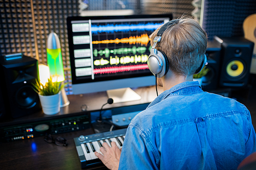 Rear view of young man in headphones looking at computer screen while making music and recording it in modern studio