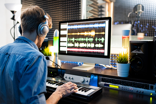 Young musician with headphones pressing keys of piano keyboard while sitting by computer monitor and working with sounds