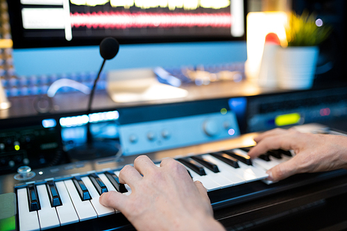 Hands of young musician pressing keys of piano keyboard in front of microphone and computer monitor while recording music