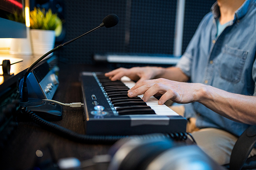 Young male musician touching keys of piano keyboard while sitting by workplace and recording new music