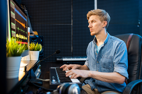 Young casual blonde man looking at computer screen while pressing keys of piano keyboard in sound recording studio