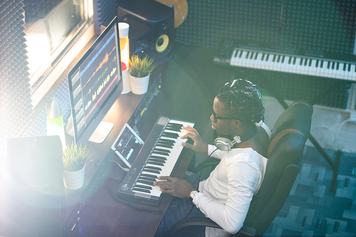 Young African musician in casualwear looking at computer screen while sitting by desk and touching keys of piano keyboard