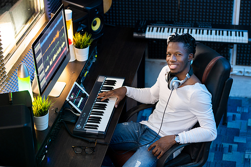 Cheerful young man in casualwear  while working over new music in front of computer monitor in studio