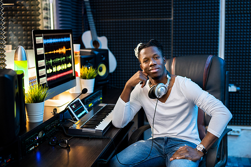 Young successful African man in casualwear sitting by workplace with computer monitor in sound record studio