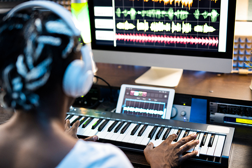 African or mixed-race young man touching keys of piano keyboard while sitting in front of computer screen and creating music