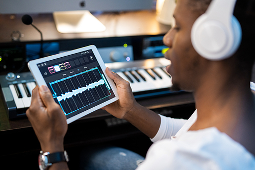African-american young man with headphones holding tablet with sound waveforms in front of himself while making music in studio