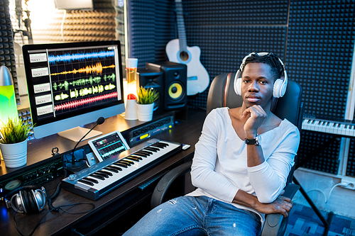 Young serious African musician in casualwear and headphones sitting in armchair by workplace in sound recording studio