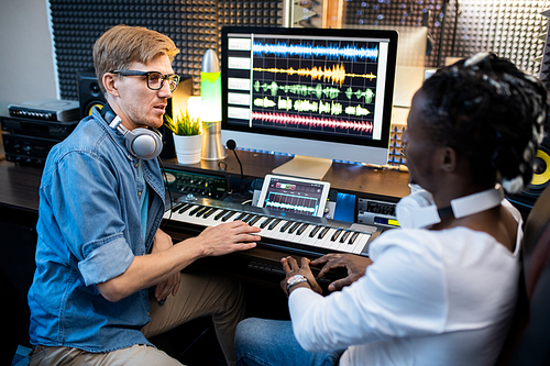 Young multi-ethnic musicians in casualwear sitting by workplace in recording studio and discussing new system of mixing sounds