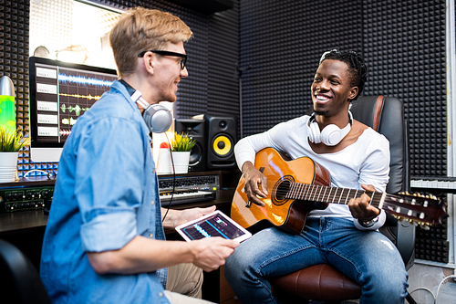 Happy young African man with guitar and his colleague sitting in modern sound record studio and working over new soundtrack
